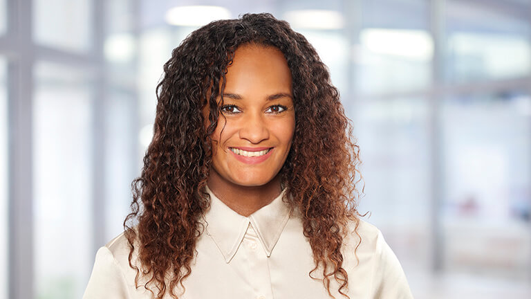 Eine Person mit lockigem Haar, die ein helles Hemd trägt, steht in einem Innenraum, möglicherweise einem Büro oder öffentlichen Gebäude.