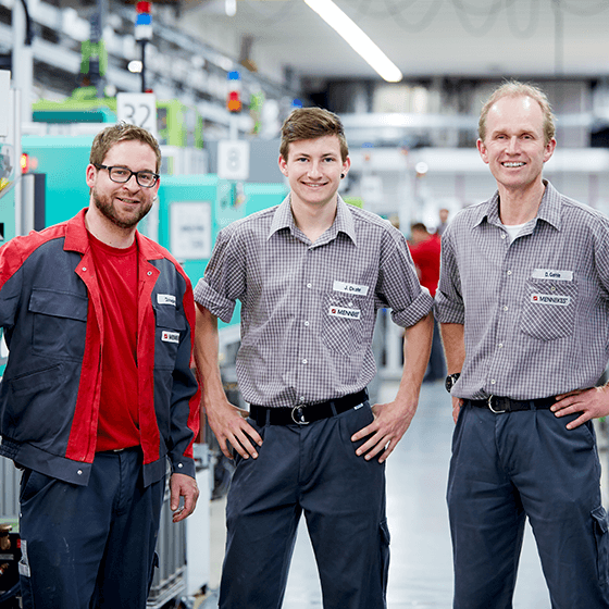 Drei Personen stehen nebeneinander in der Produktionshalle und schauen in die Kamera