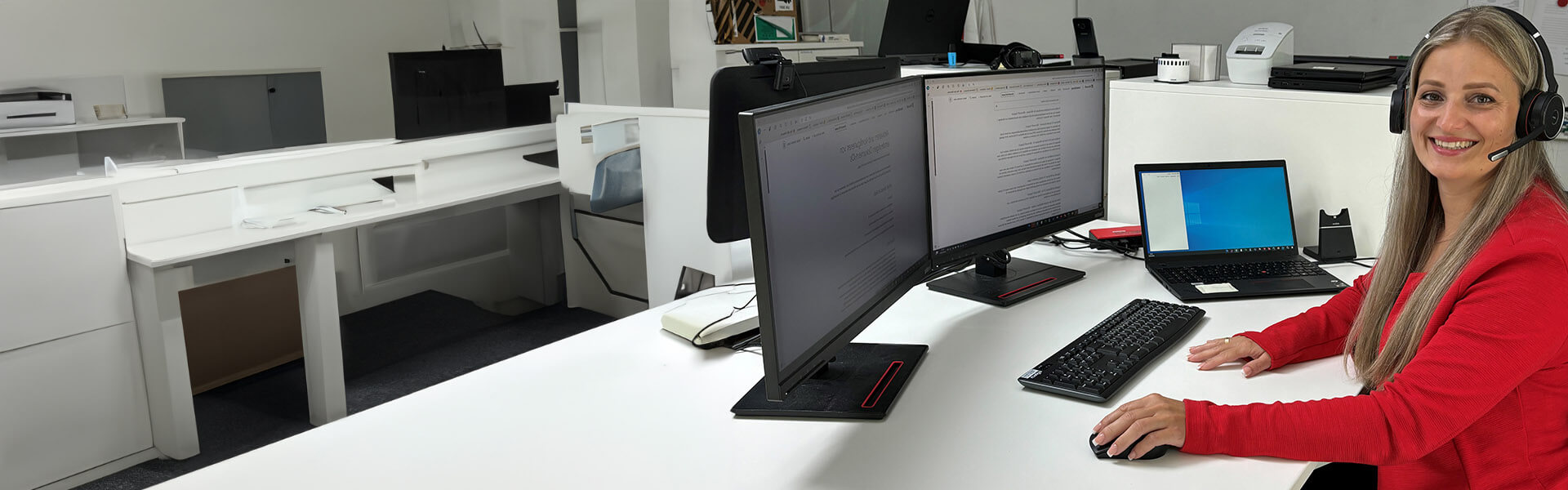 Eine Frau mit langen blonden Haaren trägt ein Headset und sitzt an einem Schreibtisch vor zwei Monitoren und einem Laptop. Sie lächelt in die Kamera, während sie eine Computermaus bedient. Der Arbeitsplatz ist modern eingerichtet, und auf den Bildschirmen sind Texte zu sehen.