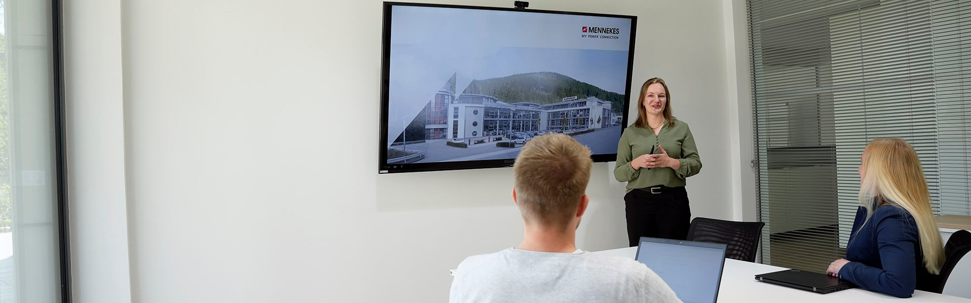 Eine Frau steht in einem Konferenzraum vor einem großen Bildschirm und hält eine Präsentation, während zwei Personen ihr zuhören und Notizen auf ihren Laptops machen.
