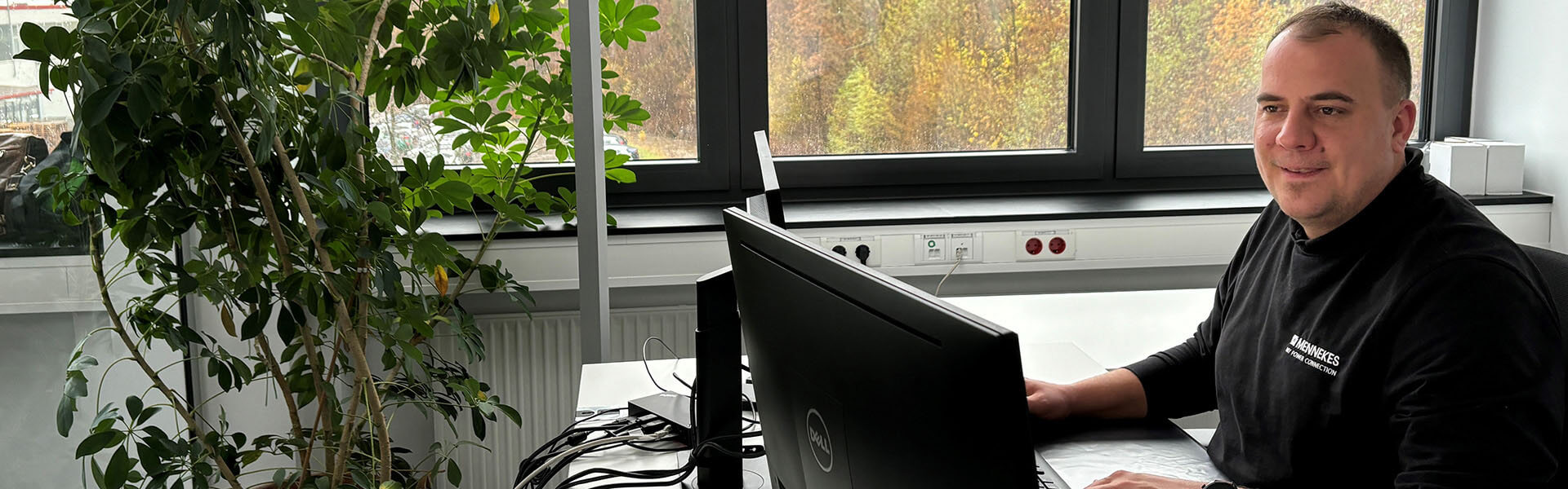 Ein Mann sitzt an einem Schreibtisch in einem modernen Büro, arbeitet am Computer und lächelt. Neben ihm steht eine große grüne Zimmerpflanze. Im Hintergrund sind Fenster zu sehen, durch die eine herbstliche Landschaft erkennbar ist