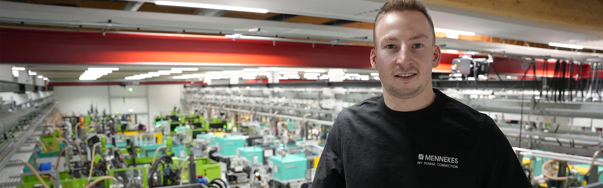 Eine männliche Person im schwarzen Pullover steht auf dem Gelände in einer Produktionshalle und lächelt in die Kamera