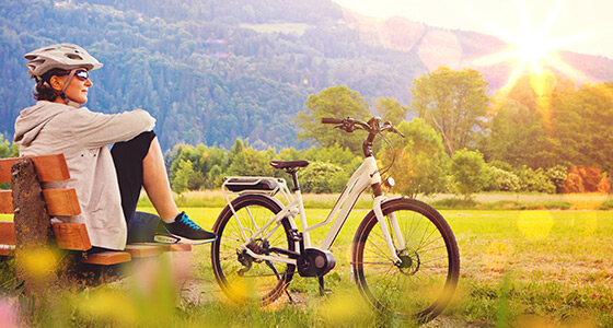 Eine Frau sitzt mit Helm und Sonnebrille auf einer Bank vor ihrem Fahrrad und schaut in den Wald rein