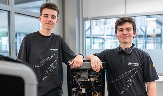 Zwei Auszubildende zum Elektroniker für Betriebstechnik stehen an einer geöffneten Steuerungseinheit in einer modernen Produktionsumgebung.