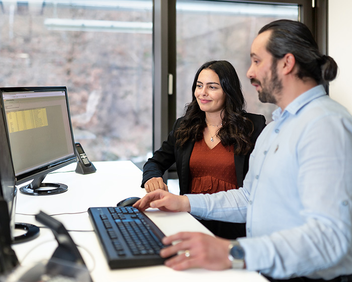 Zwei Personen stehen an einem Tisch und schauen auf einen Computerbildschirm