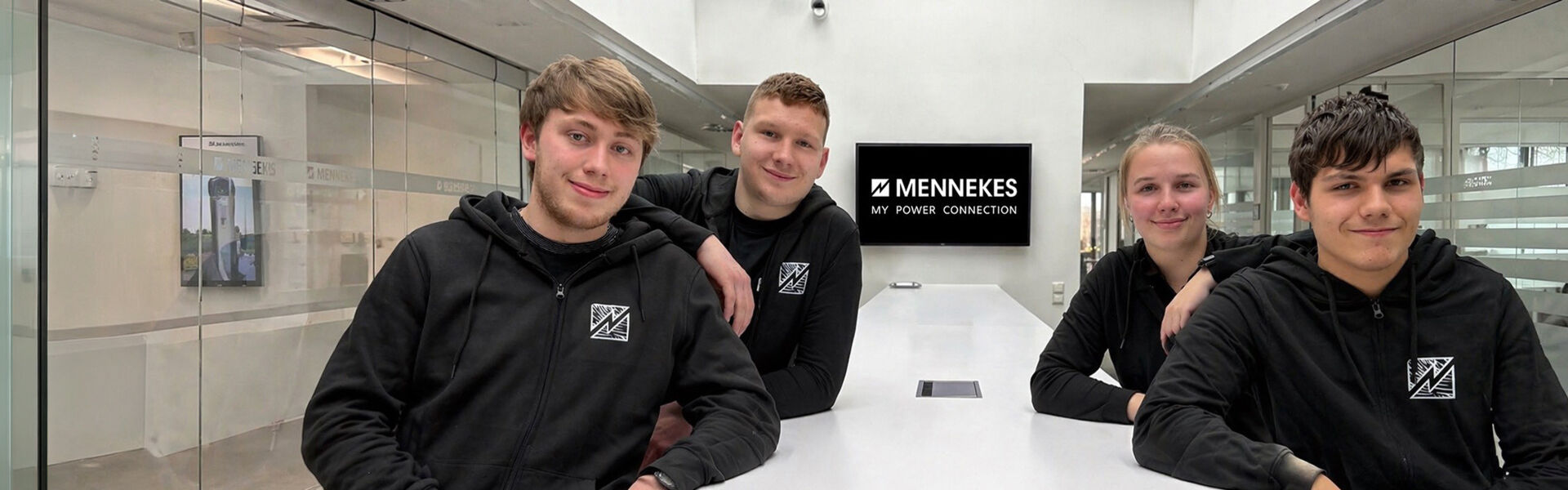 Vier Auszubildende sitzen gemeinsam an einem Tisch im modernen Büro eines Industrieunternehmens, im Hintergrund das MENNEKES Logo.