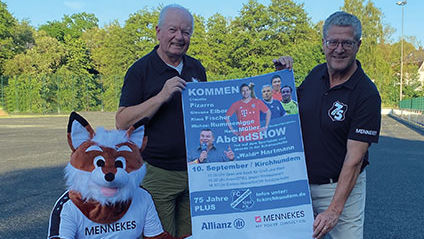 Zwei Männer halten einen Plakat zur Jubiläumsveranstaltung des FC Kirchhundem in den Händen. Daneben kniet das Vereinsmaskottchen.