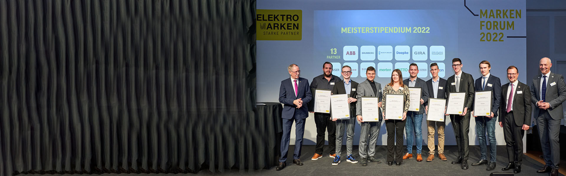 Elf Personen stehen während einer Verleihung auf dem Podium und lächeln in die Kamera. Einige von ihnen halten Urkunden in der Hand.