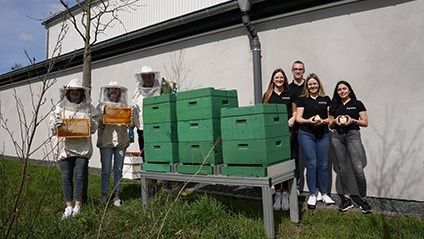 Sieben Personen stehen vor Honigräumen und halten Honiggläser in der Hand. Dabei schauen und lächeln sie in die Kamera