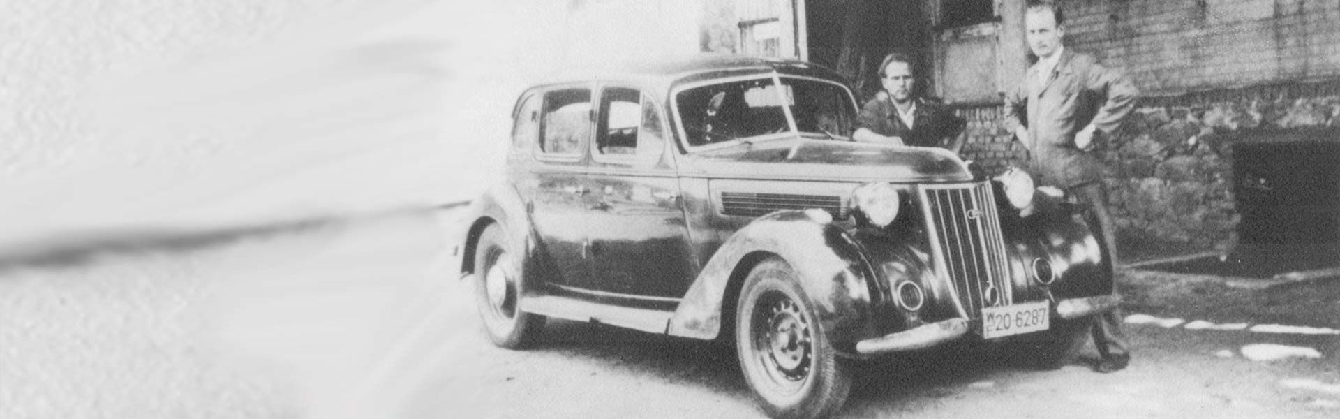 Ein historisches Bild mit zwei Männern, die versetzt hinter einem Oldtimer stehen