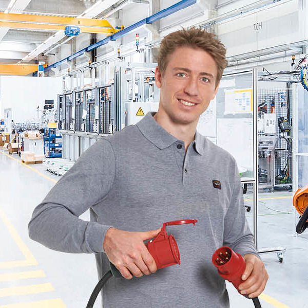 Ein Mann mit grauem Oberteil hält einen roten Stecker und eine rote Kupplung in der Hand. Im Hintergrund ist eine Produktionshalle zu sehen