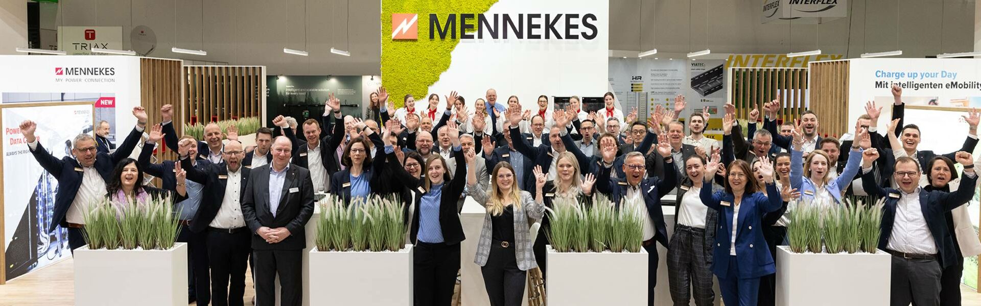 Mitarbeitende von MENNEKES stehen auf einem Messestand und heben jubelnd die Hände, im Hintergrund das MENNEKES-Logo.