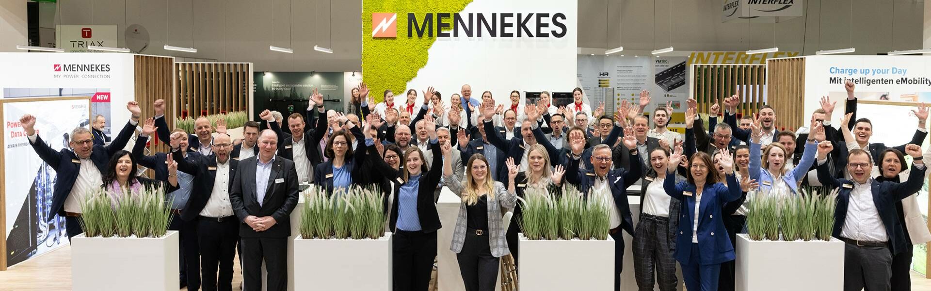 Mitarbeitende von MENNEKES stehen auf einem Messestand und heben jubelnd die Hände, im Hintergrund das MENNEKES-Logo.