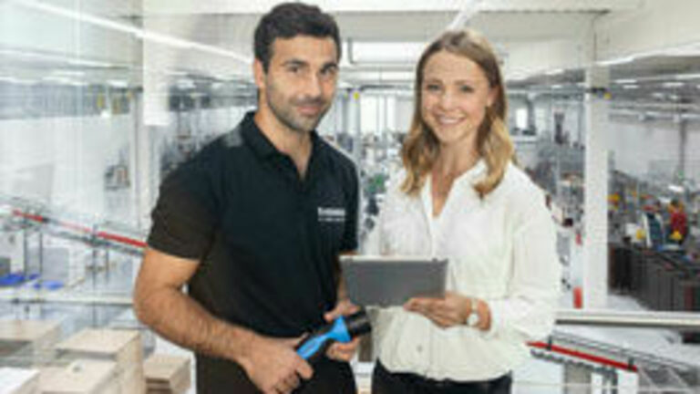 Eine Frau und ein Mann stehen in einer Produktionshalle und schauen auf ein Tablet.