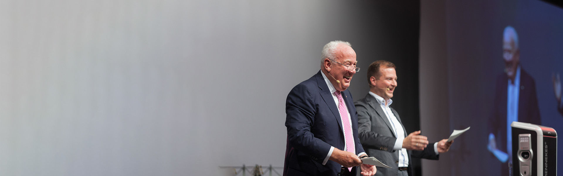 Zwei Männer stehen auf einem Podium und sprechen lächelnd zum Publikum