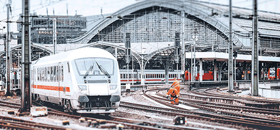 Ein weißer ICE fährt an einem Hauptbahnhof los. Im Hintergrund arbeiten Gleisarbeiter an den Gleisen