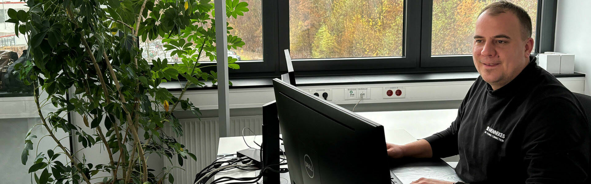 Ein Mann sitzt an einem Schreibtisch in einem modernen Büro, arbeitet am Computer und lächelt. Neben ihm steht eine große grüne Zimmerpflanze. Im Hintergrund sind Fenster zu sehen, durch die eine herbstliche Landschaft erkennbar ist